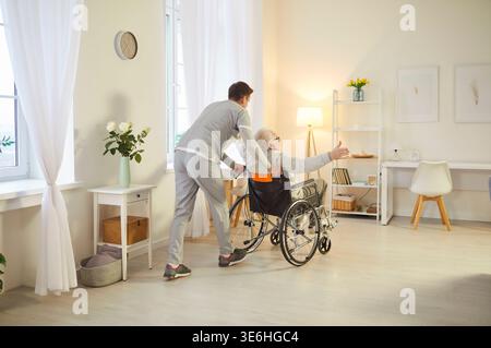 Bienveillant jeune homme soignant poussant homme âgé en fauteuil roulant qui s'amuse et apprécie la mobilité. Banque D'Images