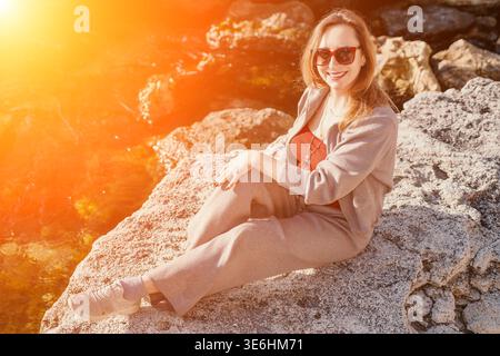 Femme côte coucher de soleil femme heureuse se trouve sur la rive rocheuse profitant de la lumière de l'heure d'or pendant ses vacances à la mer Banque D'Images