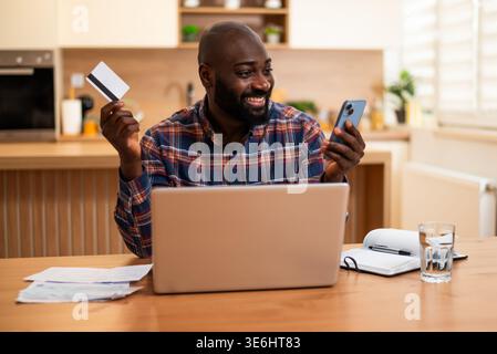 Homme noir joyeux assis à sa table de cuisine tenant une carte de crédit et un smartphone, examinant les documents financiers. Banque D'Images