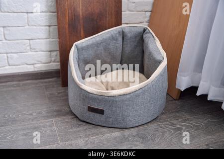 Lit d'animal de compagnie en tissu gris doux avec coussin beige moelleux à l'intérieur, positionné sur un plancher en bois sombre contre un mur de briques blanches et près de panneaux en bois et Cu clair Banque D'Images