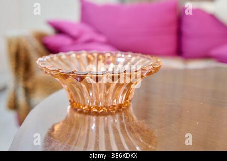 Bol en verre ambré transparent incurvé placé sur une surface de table réfléchissante lisse avec coussins roses doux flous et éléments intérieurs confortables en arrière-plan Banque D'Images