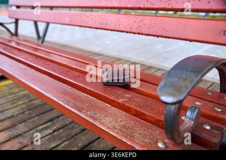 Portefeuille en cuir brun foncé vieilli reposant sur des lattes en bois humides rouges de banc de parc avec accoudoir en métal, fond de front de mer extérieur, atmosphère pluvieuse, Banque D'Images