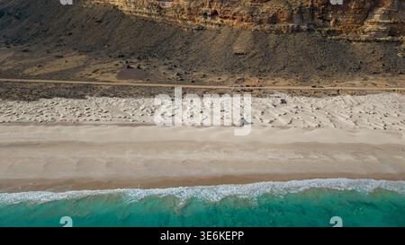 Vue aérienne des vagues turquoise de l'océan rencontrant une large plage de sable avec des motifs de dunes et des falaises désertiques accidentées en arrière-plan, terre sauvage côtière Banque D'Images