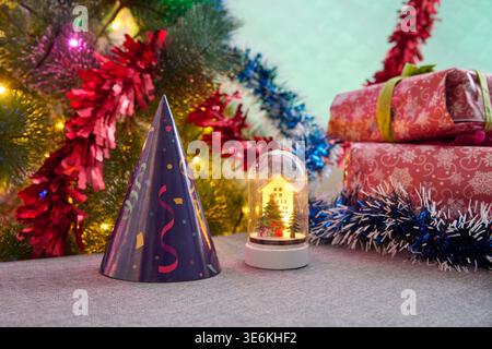 Guirlandes scintillantes colorées, chapeau de fête, boîtes cadeaux à motifs rouges empilés et boule à neige miniature brillante sur moquette souple avec dos sarcelle flou Banque D'Images