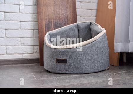 Lit d'animal de compagnie en tissu gris doux avec coussin beige moelleux à l'intérieur, positionné sur un plancher en bois sombre contre un mur de briques blanches et près de panneaux en bois et Cu clair Banque D'Images