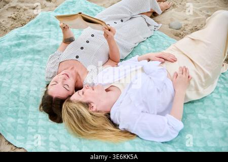 Deux femmes adultes allongées sur une couverture bleu clair sur Sandy Beach lisant paisiblement le livre ensemble sous une lumière naturelle douce. La scène transmet le calme, le loisir, et Banque D'Images