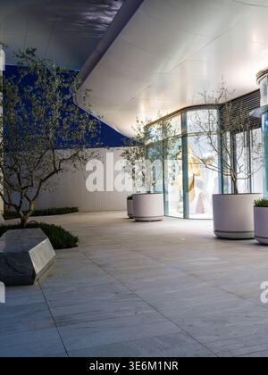 Espace architectural moderne vide avec un auvent blanc incurvé, un sol en pierre pavée et des arbres soigneusement mis en pot sous un ciel bleu foncé au crépuscule. La sce Banque D'Images