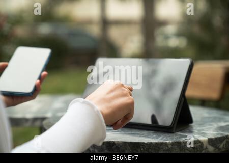 Gros plan d'une femme tenant un smartphone, concentrée sur sa tablette, dans un café en plein air. Banque D'Images