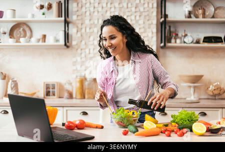 Femme prépare la salade dans la cuisine tout en regardant la vidéo sur ordinateur portable pendant la journée Banque D'Images