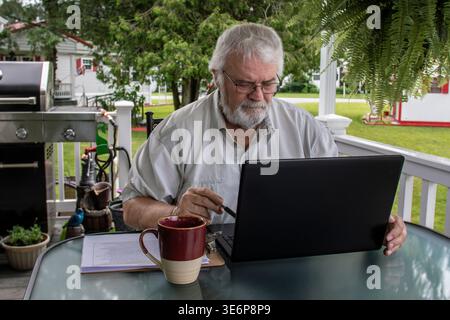 Un aîné est assis à une table dans son jardin avec un ordinateur portable ouvert devant lui. Il écrit des notes tout en tenant un stylo et apprécie une tasse de café chaud. La verdure l'entoure. Banque D'Images