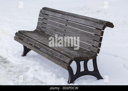 Vieux vide délavé brun à gris coloré longues lattes en bois assis banc avec des jambes en fonte noire en hiver fraîchement tombé comme la neige dans le parc public en e Banque D'Images