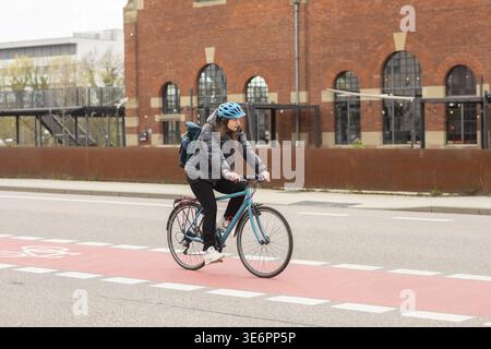Femme chevauchant un vélo sur une piste cyclable devant un grand bâtiment en briques portant un casque de vélo chevauchant un vélo dans la ville pour travailler, Freiburg im Br Banque D'Images