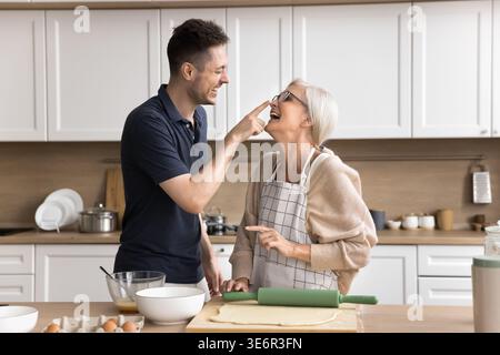 Fils adulte ludique cuisinant, s'amuser avec la mère d'âge moyen aimante dans le tablier dans la cuisine domestique confortable, riant, profiter du temps tranquille ensemble sur nous Banque D'Images
