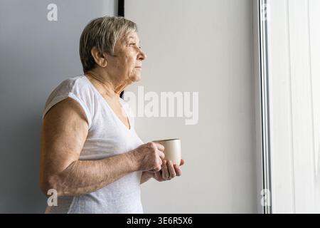 Femme senior regardant par la fenêtre lumineuse, tenant une tasse à café, pensant à la vie ou à l'avenir dans un moment calme Banque D'Images