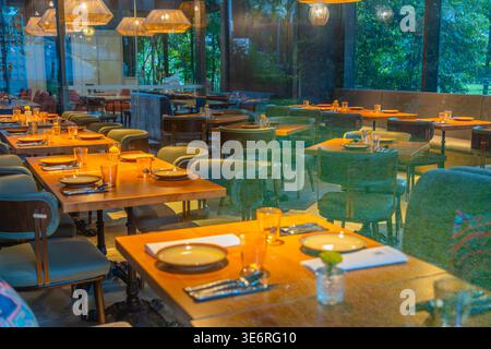 Intérieur moderne vide du restaurant avec tables en bois pour le service, chaises rembourrées vertes et grandes fenêtres avec vue sur le jardin. ECLAIRAGE chaleureux Banque D'Images