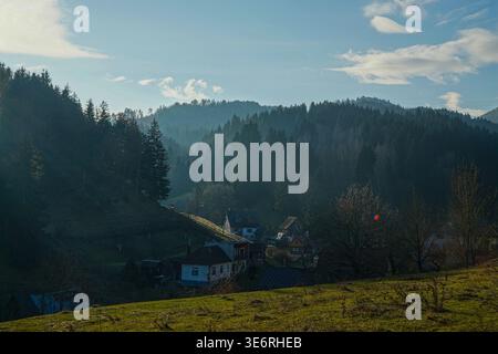 Petit village rural avec des maisons dans la vallée boisée sous la lumière douce du soleil et la brume avec des collines et des arbres en arrière-plan. Banque D'Images