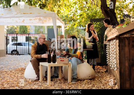 Équipe multiethnique de vendeur et de client dégustant du vin naturel fait maison, assis à table avec le vigneron à côté du stand du marché des agriculteurs. Client dégustant des produits locaux et visitant le festival gastronomique. Banque D'Images