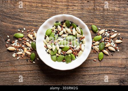 Mélange de graines dans un bol blanc sur fond en bois, vue de dessus. Texture des ingrédients alimentaires sains. Graines de citrouille, sésame noir et blanc, graines de tournesol Banque D'Images