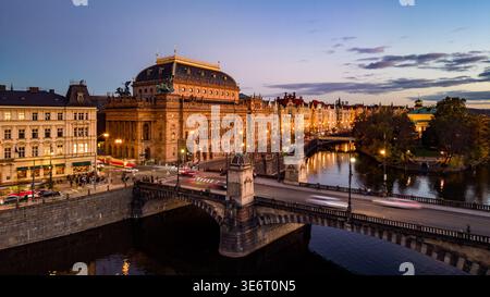 Le Théâtre National est illuminé par des lumières du soir le long du remblai à Prague. La circulation circule sur le pont voisin alors que la ville passe à la nuit. Banque D'Images