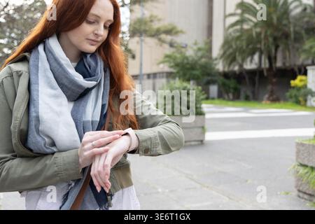 Femme aux cheveux rouges debout sur une passerelle pavée vérifiant smartwatch tout en portant une écharpe et une veste Banque D'Images