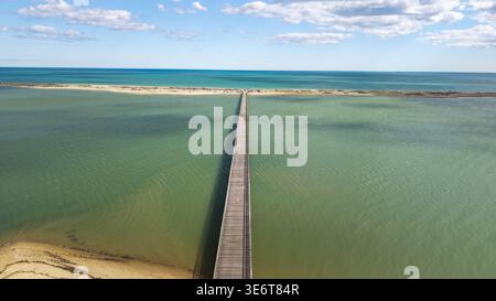 Vue aérienne d'un long pont en bois qui s'étend sur les eaux turquoises, reliant des rives sablonneuses sous un ciel de doux nuages, Duxbury, Massachuse Banque D'Images