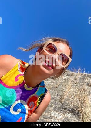 Publicité : femme souriante dans une robe vibrante penchée vers la caméra contre un ciel bleu clair d'été à Corfou. Grèce, 9 juillet 2023. Banque D'Images