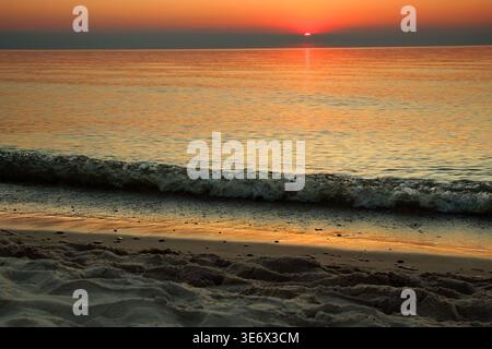 Couchant sur la mer Baltique à Ustronie Morskie, Pologne. De douces vagues s'écrasent sur la plage de sable tandis que le soleil plonge sous l'horizon, projetant une lumière orange chaude Banque D'Images