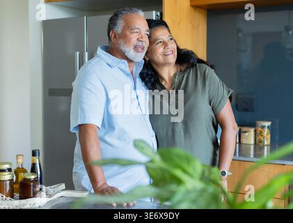 Couple senior debout dans la cuisine, penché, souriant, portant bleu clair et olive, réfrigérateur et montre Banque D'Images