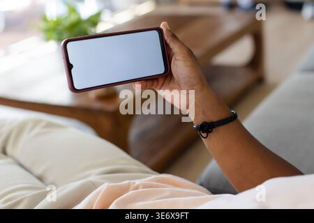 Homme afro-américain tenant smartphone horizontalement tout en s'inclinant sur un canapé de couleur claire à la maison Banque D'Images