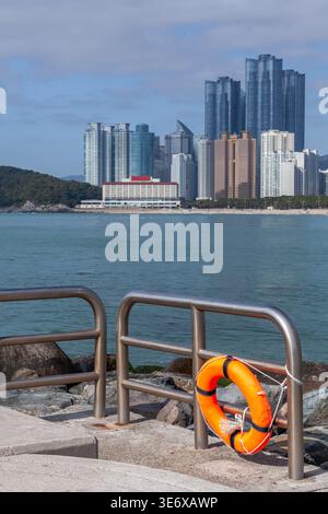 Busan, Corée du Sud. Vue sur la plage de Haeundae. Bouée de sauvetage orange attachée à une balustrade métallique sur un quai surplombant un front de mer calme et une ville moderne de grande hauteur Banque D'Images
