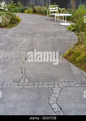 Une scène extérieure paisible avec un chemin sinueux fait de grandes dalles irrégulières de pierre grise bordées de plantes vertes et d'une chaise blanche. La scène Re Banque D'Images