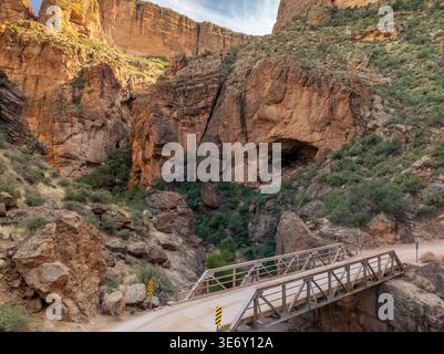 Pont métallique traversant un canyon étroit sur Apache Trail avec d'énormes falaises de roche rouge et une végétation désertique près de Phoenix Arizona sous un ciel partiellement nuageux. Banque D'Images