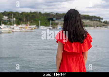 Femme en robe rouge par Marina. Vue arrière d'une femme dans une robe rouge vif debout près du front de mer calme près des bateaux et des arbres sous un ciel nuageux. Banque D'Images