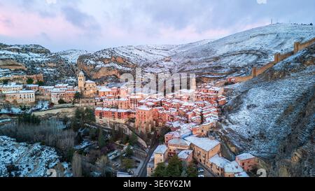 Vue aérienne spectaculaire de Albarracín sous une couverture de neige. Le village médiéval avec ses maisons roses et sa cathédrale brille dans la lumière du crépuscule, wh Banque D'Images