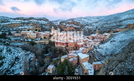 Vue aérienne spectaculaire de Albarracín sous une couverture de neige. Le village médiéval avec ses maisons roses et sa cathédrale brille dans la lumière du crépuscule, wh Banque D'Images