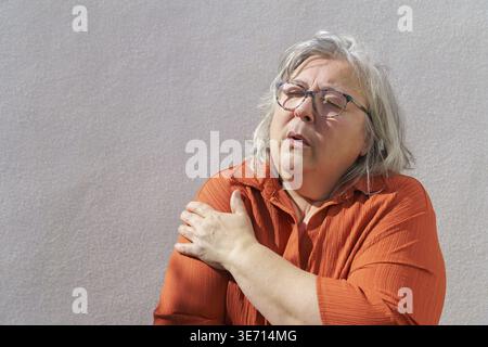 Femme âgée souffrant de douleurs chroniques à l'épaule, de symptômes d'arthrite et d'inflammation articulaire sur un fond clair, serrant son bras avec un ex Banque D'Images