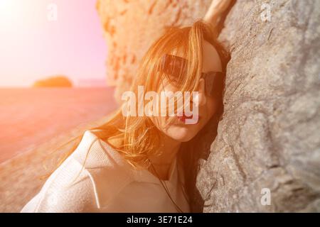 Femme lunettes de soleil côte attrayante dame portant des ombres appuyées contre un rocher sur le rivage pendant le coucher du soleil Banque D'Images