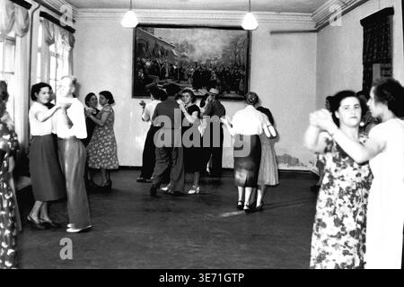 Slaviansk, 1959 ans, École technique de l'énergie. Une scène intérieure animée capturant de jeunes diplômés profitant d'une danse de célébration pendant leur soirée de remise des diplômes. Les couples dansent gracieusement sur le sol d'une salle spacieuse. En arrière-plan, une grande peinture historique dramatique est accrochée en évidence sur le mur texturé entre les luminaires. Les participants portent une élégante tenue festive du milieu du siècle, marquant la joyeuse conclusion de leurs études techniques rigoureuses avec camaraderie et musique. La vie pacifique dans le Donbass avant le conflit. Banque D'Images