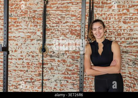 Femme adulte appuyée contre la crémaillère à la salle de gym en équipement noir avec des bandes de résistance, poulie, espace de copie. Force, fitness, entraînement, entraînement, studio, brique, Banque D'Images