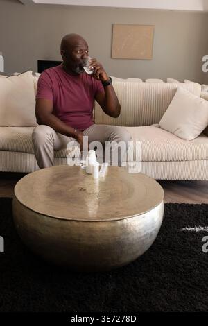 Homme chevronné marron levant le verre à la bouche tout en étant assis sur un canapé dans la salle de séjour avec casserole. Personnes âgées, gentleman, maison, intérieur, chaud, serein, Banque D'Images
