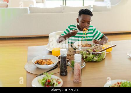 Enfant afro-américain assis dans la salle à manger mangeant des spaghettis avec une fourchette à côté d'un saladier. Pâtes, enfant, table, lumière du jour, rustique, candid, maison Banque D'Images