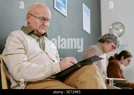 Homme principal remplissant les documents dans la salle d'attente pour la visite médicale Banque D'Images