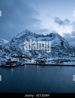 Vue aérienne des montagnes enneigées rencontrant les eaux sombres et réfléchissantes, avec de petites lumières scintillantes dans le village de Hamn, Troms, Norvège. Banque D'Images