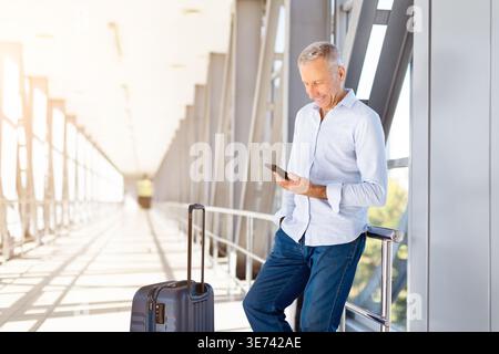 Homme mûr utilisant un smartphone dans le terminal de l'aéroport tout en se tenant debout avec des bagages Banque D'Images