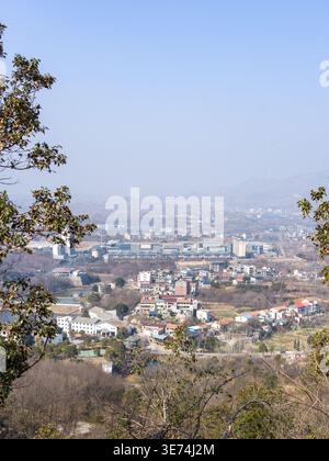 Une vue panoramique surélevée depuis une colline boisée surplombant une ville lointaine ou une ville située dans une vallée, encadrée par des arbres sous un ciel bleu brumeux. La scène R Banque D'Images
