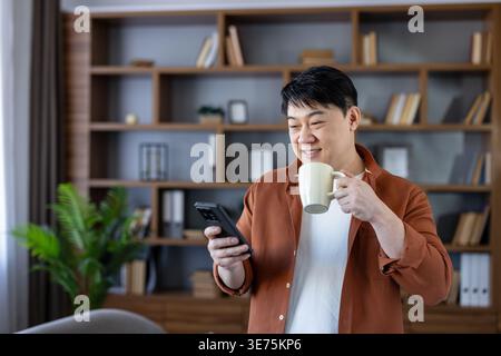 Homme asiatique souriant tenant une tasse à café tout en parcourant le contenu sur un smartphone, profitant d'un moment de détente dans un bureau à domicile ou un salon avec une étagère en arrière-plan Banque D'Images