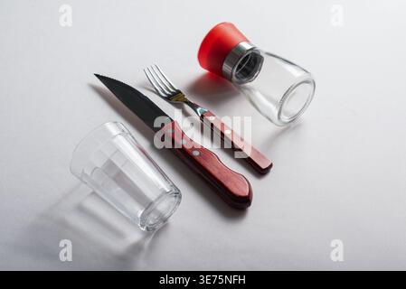 Composition minimaliste top-down avec couteau, fourchette, verre et salière sur une surface légère. Un éclairage clair et lumineux accentue les textures de verre et de métal Banque D'Images