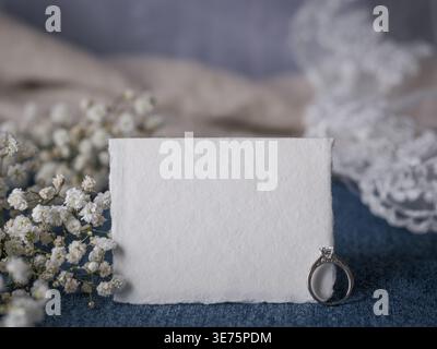 Carte en papier vierge faite à la main avec bague de fiançailles et fleurs disposées sur denim créant une invitation de mariage occidental rustique ou un message de proposition romantique Banque D'Images