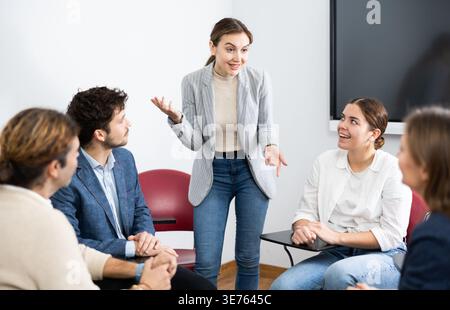 Une entraîneure professionnelle se tenant près du tableau interactif et menant la formation de différentes personnes rassemblées en cercle dans l'auditorium Banque D'Images