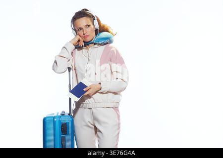 Une femme dans un survêtement rose semble inquiète tout en tenant son passeport, préparée pour le voyage avec un oreiller de cou et une valise sur un fond blanc Banque D'Images